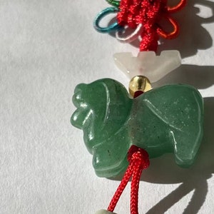 K&ouml;nnte beinhalten: Ein gr&uuml;ner Jade-Tier-Anh&auml;nger mit roter und wei&szlig;er Schnur und Perlen. Der Anh&auml;nger ist mit einem Knoten gebunden und hat eine wei&szlig;e Perle dar&uuml;ber.