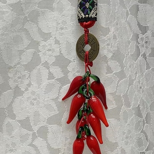 May include: A red and green glass chilli pepper charm with a red string and a brass coin with Chinese characters. The charm is hanging from a red and black fabric tassel.