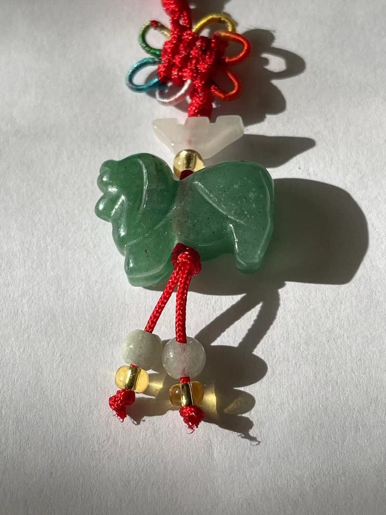 K&ouml;nnte beinhalten: Ein gr&uuml;ner Jade-Tier-Anh&auml;nger mit roter und wei&szlig;er Schnur und Perlen. Der Anh&auml;nger ist mit einem Knoten gebunden und hat eine wei&szlig;e Perle dar&uuml;ber.