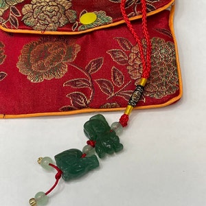 Può includere: Una pochette in tessuto rosso con ricami floreali dorati e un ciondolo in giada verde con un fiocco rosso. Il ciondolo presenta due elefanten in giada.