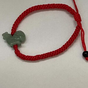 May include: Red braided bracelet featuring a light green rooster charm. The adjustable bracelet has black beads at the ends of the drawstrings. The rooster charm is a light green color.