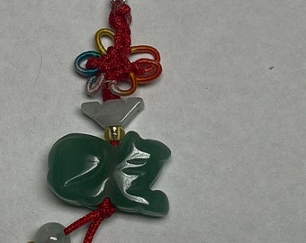 May include: A green jade charm in the shape of a Chinese character, attached to a red string with a silver clasp and a multi-colored knot. The charm is hanging from two green jade beads.