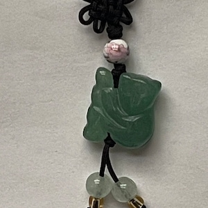 May include: A green jade pendant in the shape of a sitting Buddha, with a black cord and beads. The pendant is attached to a black cord with a knot at the top and a pink and white bead.