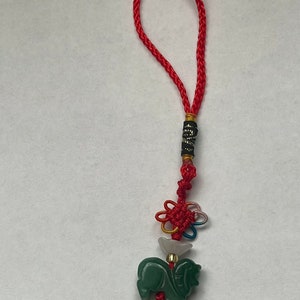 K&ouml;nnte beinhalten: Ein gr&uuml;ner Jade-Pferdeanh&auml;nger mit einer roten und wei&szlig;en Schnur und einem mehrfarbigen Knoten. Der Anh&auml;nger ist an einer roten Schnur mit einer schwarzen und goldenen Perle befestigt.