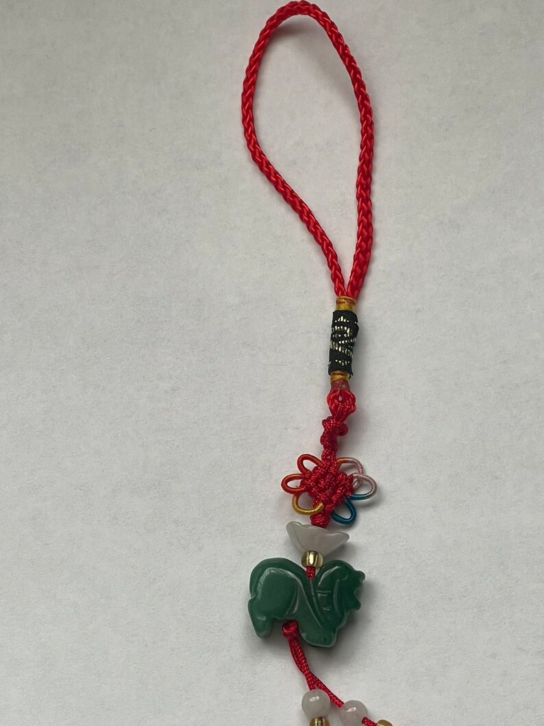 K&ouml;nnte beinhalten: Ein gr&uuml;ner Jade-Pferdeanh&auml;nger mit einer roten und wei&szlig;en Schnur und einem mehrfarbigen Knoten. Der Anh&auml;nger ist an einer roten Schnur mit einer schwarzen und goldenen Perle befestigt.