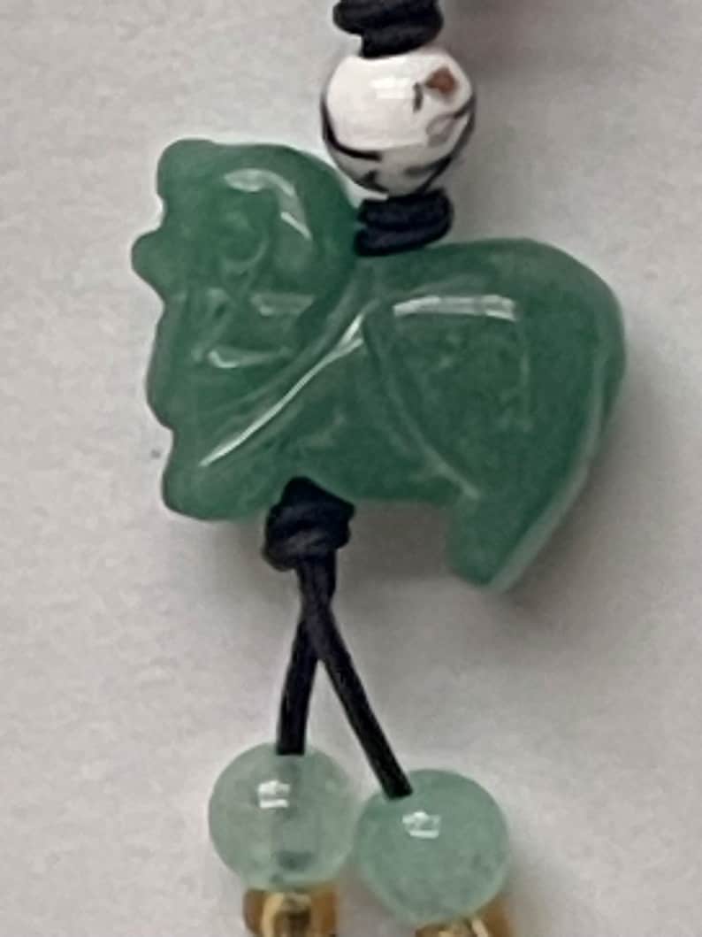 K&ouml;nnte beinhalten: Ein gr&uuml;ner Jade-Anh&auml;nger in Form eines Fabelwesens, mit einer schwarzen Schnur und wei&szlig;en und braunen Perlenakzenten.