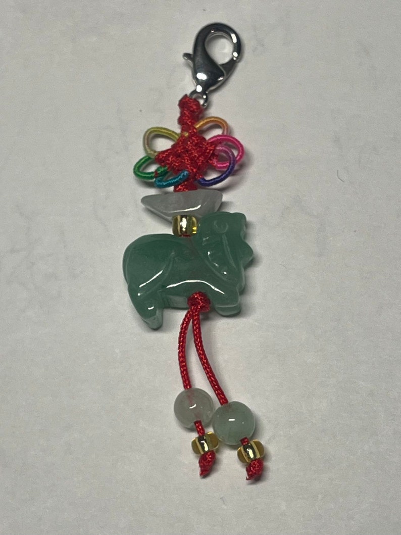 K&ouml;nnte beinhalten: Gr&uuml;ner Jade-Ochsen-Anh&auml;nger mit roten und goldenen Akzenten. Der Ochse ist an einer roten Schnur mit einem silbernen Clip und einem mehrfarbigen Knoten befestigt.