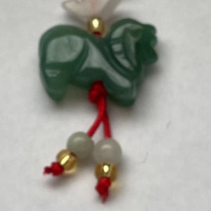 K&ouml;nnte beinhalten: Ein gr&uuml;ner Jade-Anh&auml;nger in Form eines Pferdes, der an einer roten Schnur mit wei&szlig;en und goldenen Perlen h&auml;ngt. Der Anh&auml;nger ist Teil einer gr&ouml;&szlig;eren Schnur mit Anh&auml;ngern in Rot, Wei&szlig; und Blau.