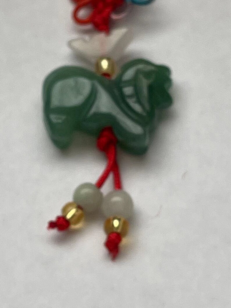 K&ouml;nnte beinhalten: Ein gr&uuml;ner Jade-Anh&auml;nger in Form eines Pferdes, der an einer roten Schnur mit wei&szlig;en und goldenen Perlen h&auml;ngt. Der Anh&auml;nger ist Teil einer gr&ouml;&szlig;eren Schnur mit Anh&auml;ngern in Rot, Wei&szlig; und Blau.