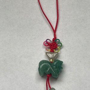 K&ouml;nnte beinhalten: Ein gr&uuml;ner Jade-Anh&auml;nger in Form eines Hundes mit einer roten Schnur und einem mehrfarbigen Knoten. Der Anh&auml;nger ist an einer roten Schnur mit Perlen befestigt.