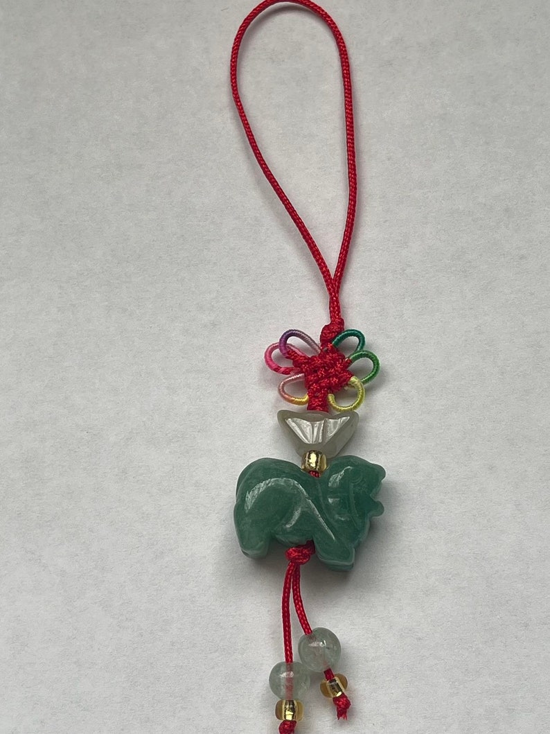 K&ouml;nnte beinhalten: Ein gr&uuml;ner Jade-Anh&auml;nger in Form eines Hundes mit einer roten Schnur und einem mehrfarbigen Knoten. Der Anh&auml;nger ist an einer roten Schnur mit Perlen befestigt.