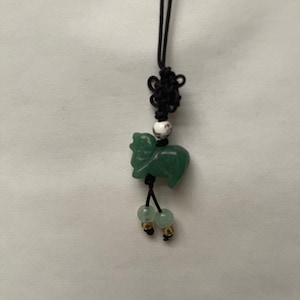 K&ouml;nnte beinhalten: Ein gr&uuml;ner Jade-Anh&auml;nger in Form eines Schafs, der an einem schwarzen Band mit einer wei&szlig;en Perle und zwei gr&uuml;nen Perlen mit goldenen Akzenten h&auml;ngt.