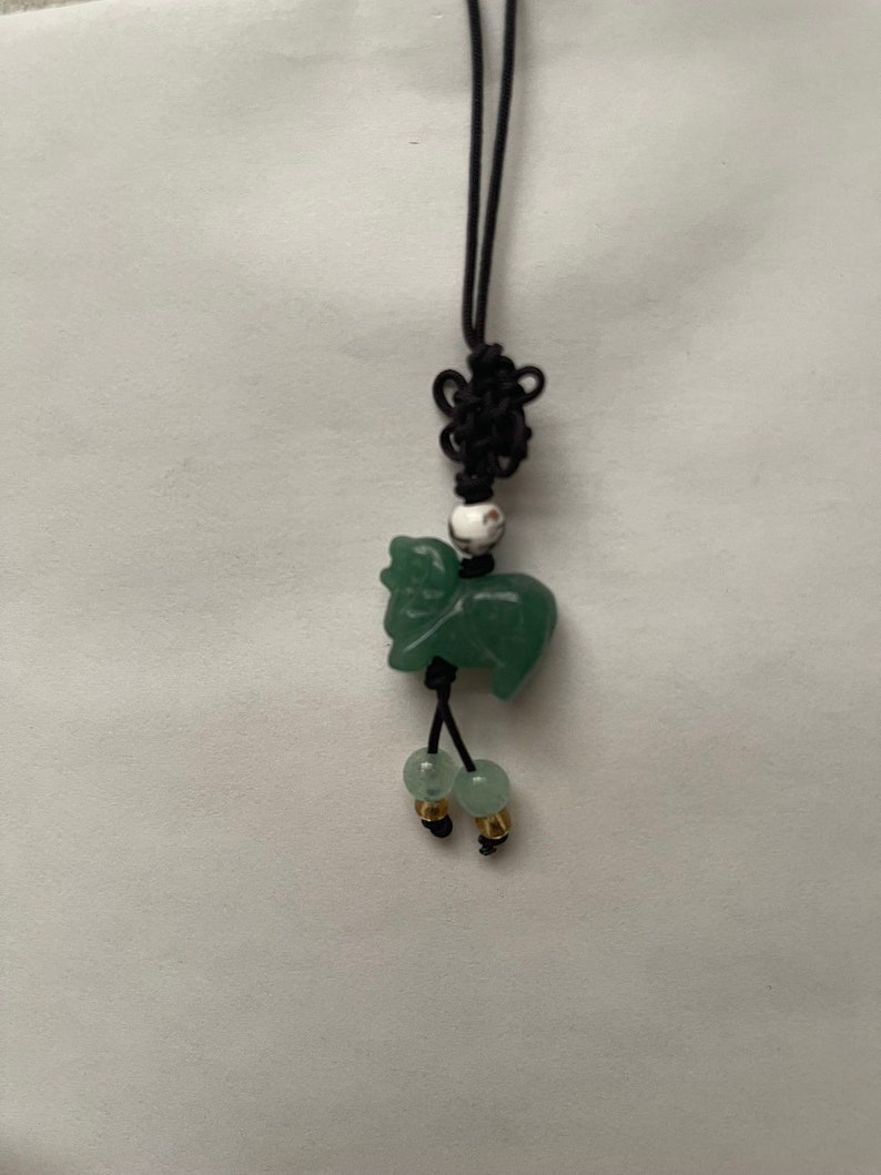 K&ouml;nnte beinhalten: Ein gr&uuml;ner Jade-Anh&auml;nger in Form eines Schafs, der an einem schwarzen Band mit einer wei&szlig;en Perle und zwei gr&uuml;nen Perlen mit goldenen Akzenten h&auml;ngt.