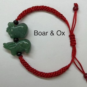 May include: A red braided bracelet featuring two green jade animal carvings, a boar and an ox, connected by black beads. The text "Boar & Ox" is displayed on the image.