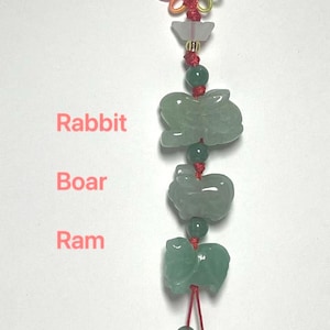 May include: A decorative hanging ornament featuring three carved green stone animal figures labelled Rabbit, Boar, and Ram. The figures are strung on a red cord with small green beads, a translucent white accent piece, and a colourful knotted top loop.