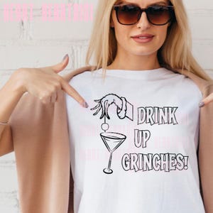 May include: White t-shirt with a black graphic of a hand, martini glass, and the text "DRINK UP GRINCHES!". The design is centered on the front of the shirt.
