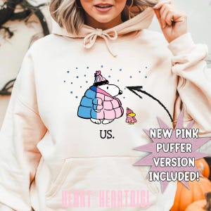 Puede incluir: Sudadera con capucha color crema con un personaje de dibujos animados Snoopy que lleva una chaqueta y un gorro acolchados azules y rosas. El texto "US." está debajo de la imagen. También se muestra un personaje más pequeño con un atuendo rosa.