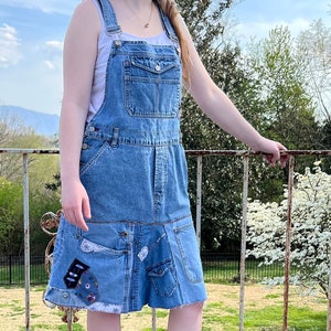 May include: A woman wearing a light blue denim overall dress with patches and a wide-brimmed straw hat. She is standing on a wooden deck with a metal railing in the background.