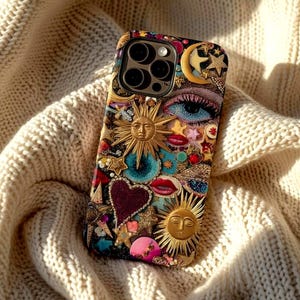 Könnte beinhalten: Dekorative Handyhülle mit einer Collage aus himmlischen und romantischen Elementen. Die Hülle enthält eine goldene Sonne, einen Mond mit Sternen, ein Herz und ein Auge. Die Hülle liegt auf einer cremefarbenen Strickdecke.