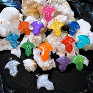 Op de afbeelding: Een verzameling kleurrijke hars hangers in diverse tinten, waaronder roze, oranje en blauw. Elke hanger heeft een uniek wit sterrenbeeld ontwerp en een zilveren of koperen oogje om op te hangen. De hangers worden gepresenteerd op een bed van bergkristal.