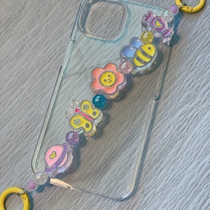 Op de afbeelding: Een transparante telefoonhoes met een kleurrijk bedeltjeskoord. Het koord is versierd met verschillende bedeltjes, waaronder een bloem, een vlinder, een bij, een snoepje en een planeet. De bedeltjes zijn allemaal van transparant plastic en hebben een glanzende afwerking.