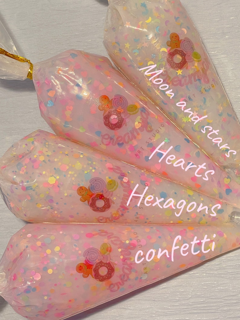May include: Four clear plastic bags filled with colorful confetti. The bags are labeled with the words "Moon and stars", "Hearts", "Hexagons", and "confetti". Each bag has a gold twist tie at the top and a logo that says "Creamy Art Studio".