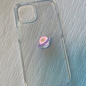 Op de afbeelding: Een transparante telefoonhoes met een roze en paarse planeet sticker met een geel hartje.