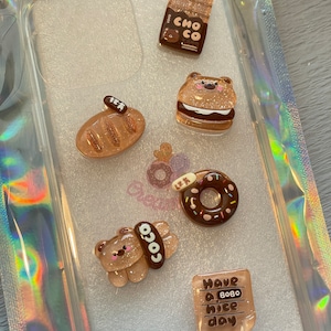 May include: A clear phone case with several decorative resin charms. The charms include a chocolate jar, a bear-shaped sandwich, a loaf of bread, a donut, a bear with a "coco" label, and a small square with the text "Have a bobo nice day."