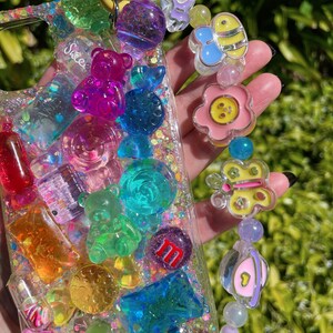 Op de afbeelding: Een transparante telefoonhoes met een kleurrijk, glitterend ontwerp met gummyberen, snoepjes en andere zoete lekkernijen. De hoes heeft een gele band met een bedel eraan, met een bloem, vlinder en bij.