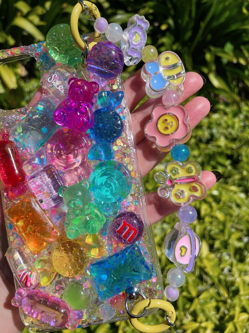 Op de afbeelding: Een transparante telefoonhoes met een kleurrijk, glitterend ontwerp met gummyberen, snoepjes en andere zoete lekkernijen. De hoes heeft een gele band met een bedel eraan, met een bloem, vlinder en bij.