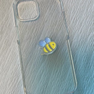 Op de afbeelding: Een transparante telefoonhoes met een geel en zwart bijenontwerp. De bij heeft blauwe vleugels.