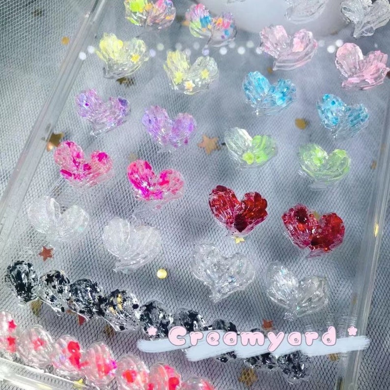 May include: A collection of heart-shaped embellishments in various colors, including pink, red, blue, and clear, with glitter accents. The hearts are arranged on a clear surface, creating a decorative and sparkling effect. Some hearts have star-shaped details.