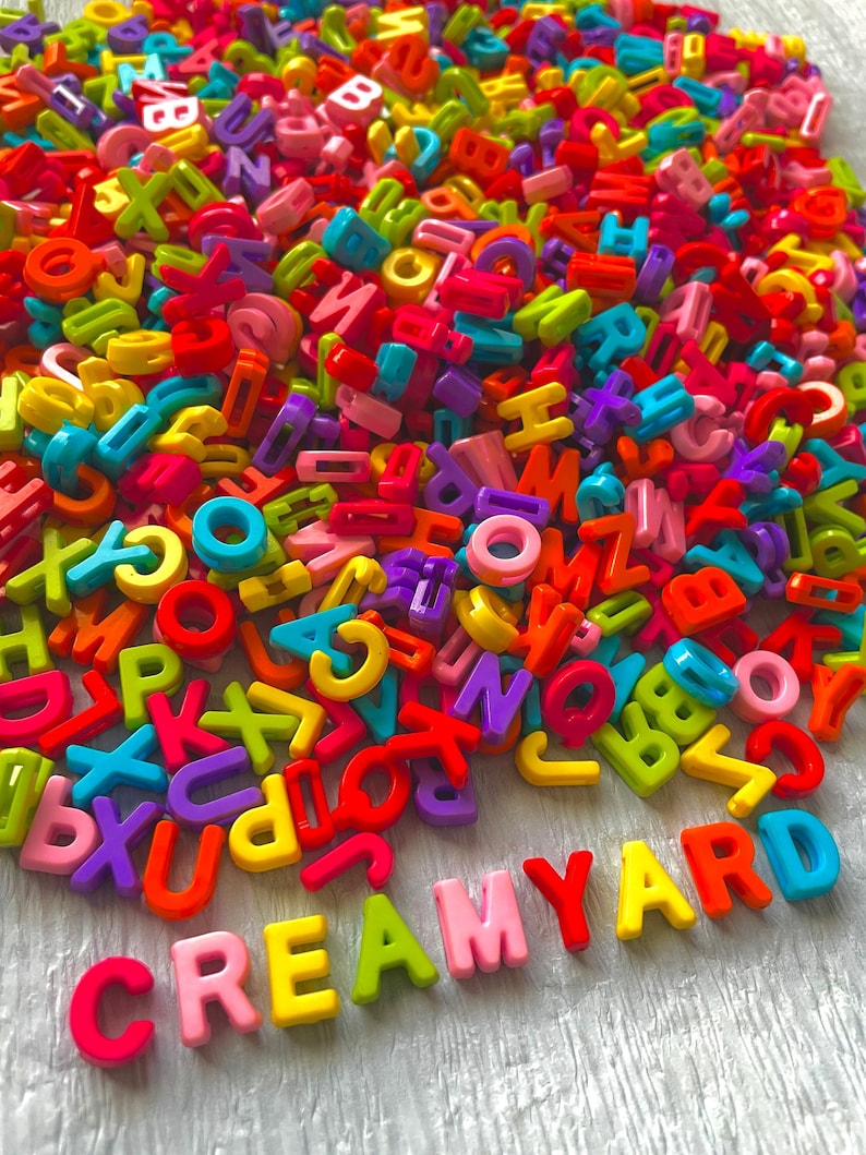 Może przedstawiać: Stosik kolorowych plastikowych liter układających się w słowo "CREAMYARD" na białym drewnianym tle.