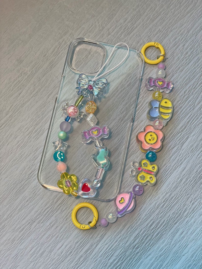 Op de afbeelding: Een transparante telefoonhoes met een kleurrijk kralen telefoonkoord. Het koord heeft bedels in de vorm van een vlinder, een bij, een bloem, een snoepje, een hart en een planeet. Het koord is bevestigd aan de telefoonhoes met een gele metalen ring.
