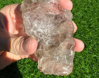 May include: A large, clear quartz crystal cluster with a light pink hue. The crystal has many points and facets, and it is translucent. The crystal is sitting on a green surface.