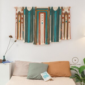 May include: A macrame wall hanging with a mix of white, teal, orange, and brown yarn. The hanging is made up of several sections, each with a different pattern. The hanging is mounted on a white wall above a bed with pillows and a book.