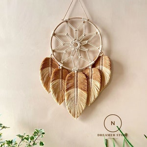May include: A beige and brown macrame dream catcher with a circular woven design and hanging feather accents. The dream catcher is suspended by a string against a neutral wall. The bottom of the image shows green plants.