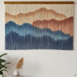 Peut inclure: Une grande tenture murale représentant un paysage de montagne. L'œuvre utilise un dégradé de couleurs, passant du beige au marron et au bleu. La pièce est montée sur une barre en bois et est suspendue à un mur blanc.