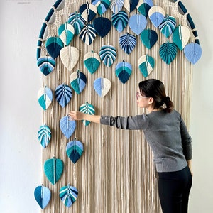 May include: A large macrame wall hanging with a circular frame and a design of blue and green leaves. The wall hanging is made of natural cotton cord and is decorated with a variety of blue and green yarn leaves.