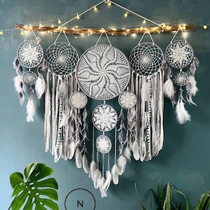 May include: A set of six grey and white dreamcatchers with feathers hanging from a wooden branch. The dreamcatchers are arranged in a cascading pattern, with the largest dreamcatcher at the top and the smallest dreamcatcher at the bottom. The dreamcatchers are decorated with white crocheted designs.