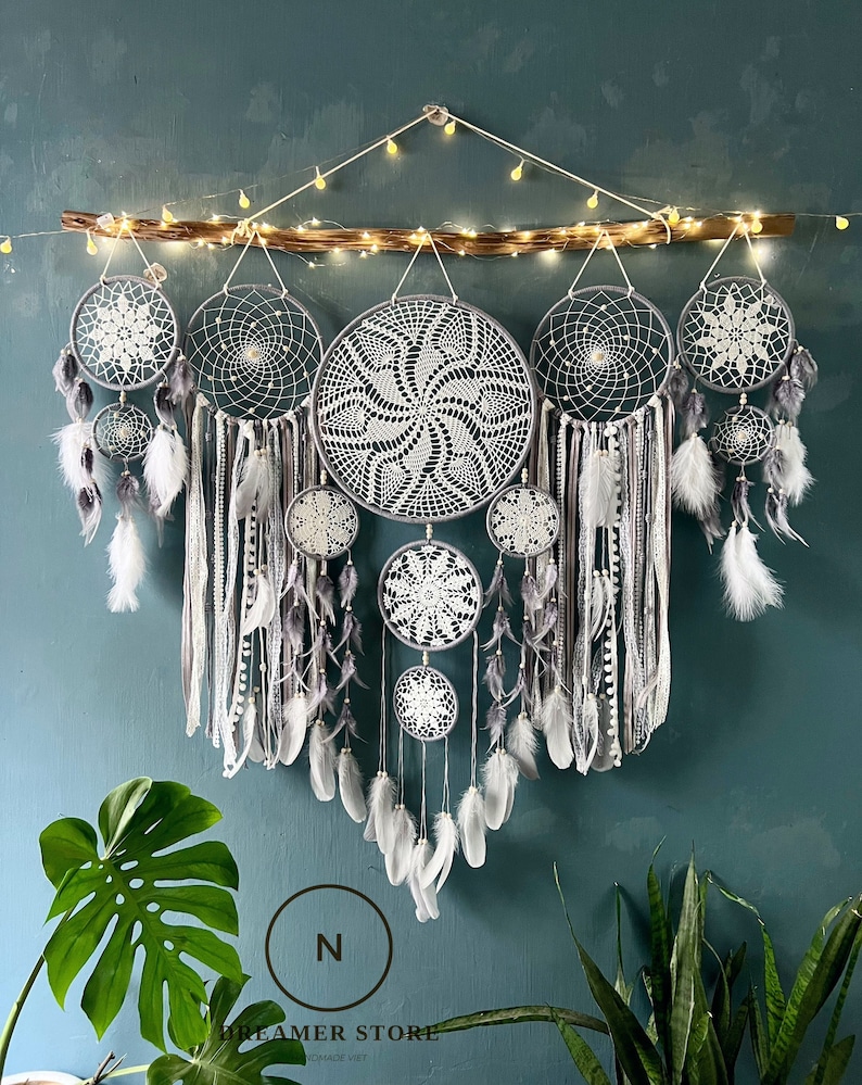 May include: A set of six grey and white dreamcatchers with feathers hanging from a wooden branch. The dreamcatchers are arranged in a cascading pattern, with the largest dreamcatcher at the top and the smallest dreamcatcher at the bottom. The dreamcatchers are decorated with white crocheted designs.