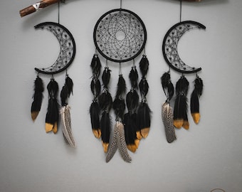 Puede incluir: Tres atrapasueños negros con plumas colgando de una rama de madera. Los atrapasueños tienen forma de círculo y de media luna. Las plumas son negras y doradas.