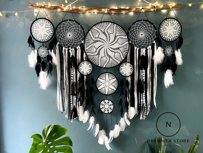 May include: A set of seven black and white dreamcatchers with feathers hanging from the bottom. The dreamcatchers are arranged in a row, with the largest one in the centre. The dreamcatchers are made of macrame and have a circular shape. The feathers are white and black.