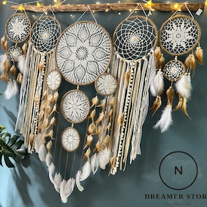 May include: A collection of seven white and brown dreamcatchers with feathers hanging from the bottom. The dreamcatchers are arranged in a row and are hanging from a wooden branch. The background is a light blue wall.