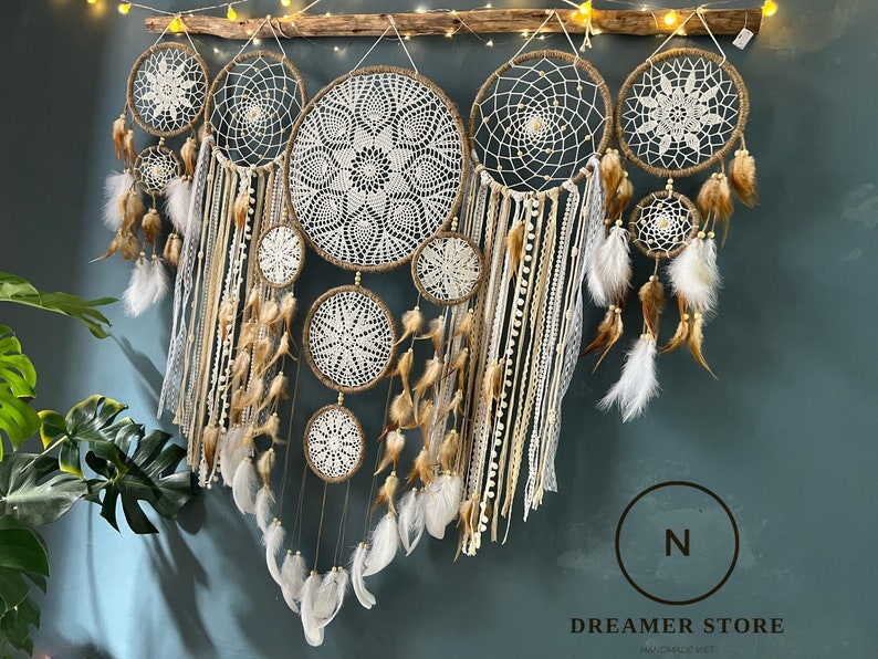 May include: A collection of seven white and brown dreamcatchers with feathers hanging from the bottom. The dreamcatchers are arranged in a row and are hanging from a wooden branch. The background is a light blue wall.