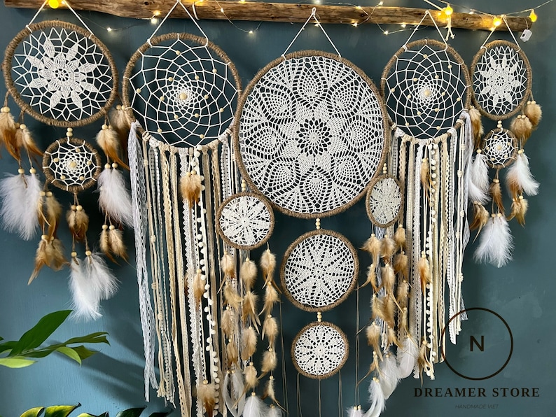 May include: A set of seven white and brown dreamcatchers with intricate crocheted designs and feathers hanging from the bottom. The dreamcatchers are arranged in a row against a blue wall.