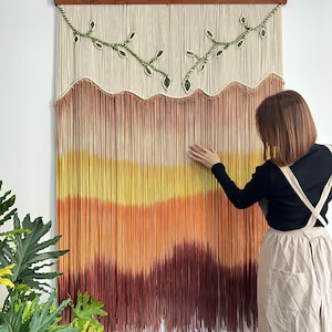 May include: A large, rectangular macrame wall hanging with a wooden top. The design features a gradient of colors, transitioning from cream to yellow, orange, and deep red. Green leaf accents are present.