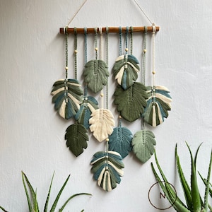 May include: A wall hanging made of macrame with a wooden dowel at the top. The macrame is in the shape of leaves in shades of green, blue, and white. The wall hanging is hanging on a white wall with a green plant in the bottom right corner. The text "DREAMER STO" is visible in the bottom right corner.