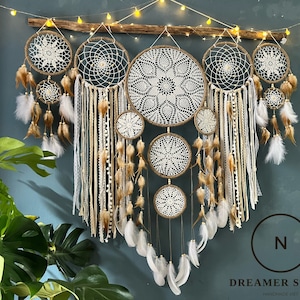 May include: A set of six macrame dreamcatchers with white and brown feathers hanging from a wooden branch. The dreamcatchers are made with white and brown yarn and have a variety of patterns. The dreamcatchers are hanging against a dark blue wall.