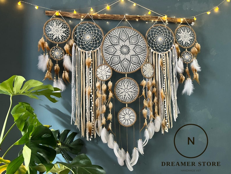 May include: A set of six macrame dreamcatchers with white and brown feathers hanging from a wooden branch. The dreamcatchers are made with white and brown yarn and have a variety of patterns. The dreamcatchers are hanging against a dark blue wall.